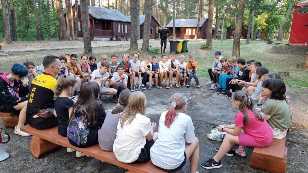 Dzieci siedzące w kręgu na ławach z kłód między drewnianymi domkami ośrodka Waszeta